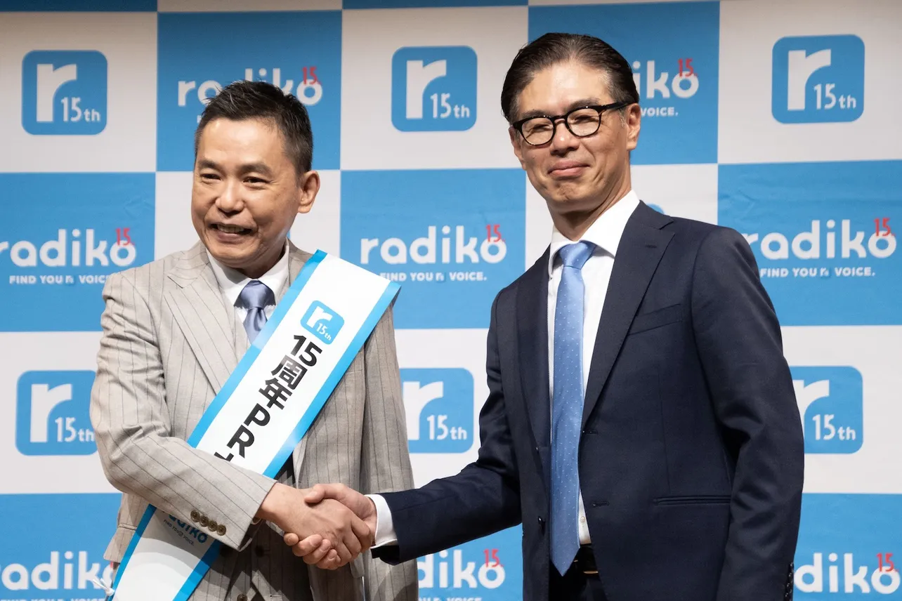 【写真】株式会社radiko 代表取締役社長・池田卓生氏よりタスキを受け取り、ガッチリ握手する太田光