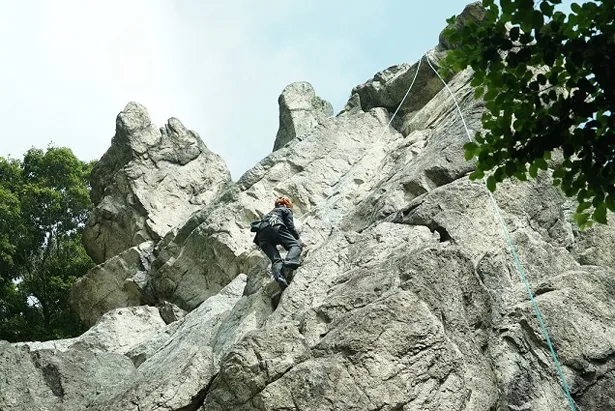 【写真を見る】スーツ姿で高さ20m以上の岩場をスーツ姿で登っていく“違和感ありまくり”な光景