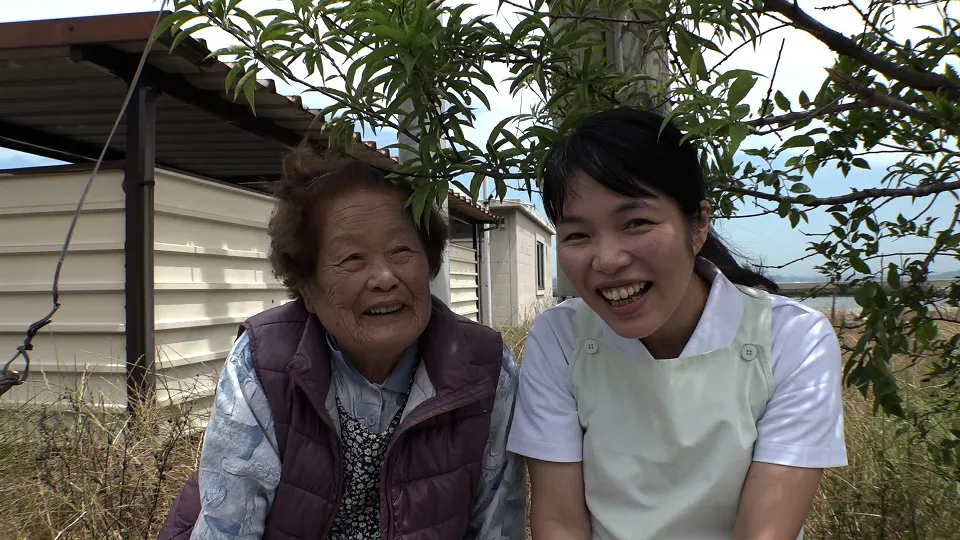 【写真を見る】島のお年寄りの隣で優しい笑顔を見せる小澤詠子さん