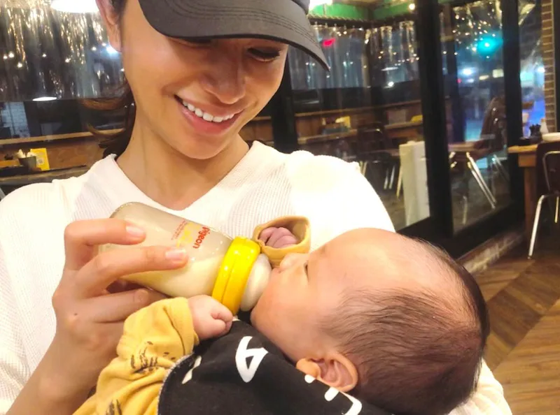【写真を見る】妹の高橋ユウの息子を笑顔で抱っこしながら、ミルクをあげる高橋メアリージュン