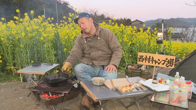 菜の花畑をバックにキャンプ飯を作る西村
