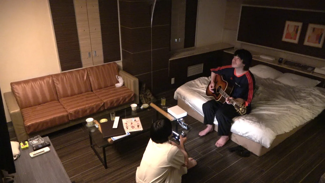 【写真を見る】ベッドに腰掛け弾き語りを披露する峯田和伸とその様子を撮影する松本穂香