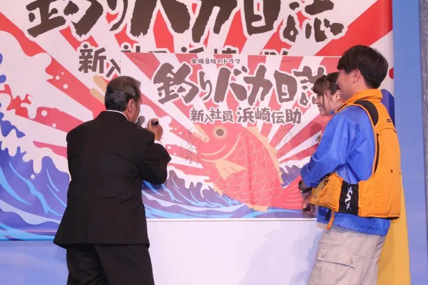 会見に用意された大漁旗にサインとイラストを入れる西田とそれを見守る濱田＆広瀬